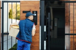 A delivery person holds a package while speaking into an intercom entry system mounted on a modern wooden gate. This residential access control setup is part of a video intercom solution, ensuring secure, contactless visitor communication in a smart building security environment.